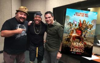 Three men with their arms linked, smiling, next to a promotional poster for a cartoon