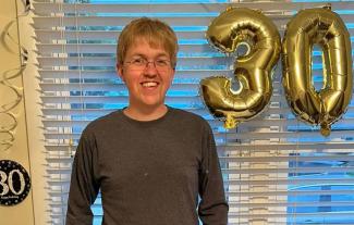 A man smiling, next to balloons in the shape of the number 30