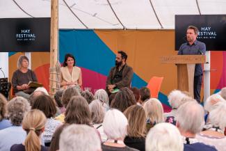 Four people on stage at an event. Two women and a man are seated and a man is standing at a lectern speaking to the audience