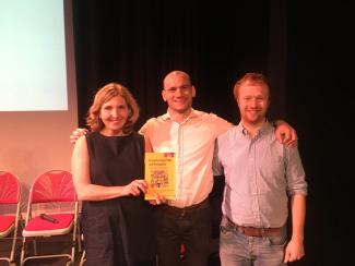 A woman and two men standing with their arms around each other and smiling. The woman is holding up a book