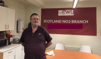 A man in a kitchen in front of a sign saying "CWU - the communications union" and "Scotland No.2 branch"