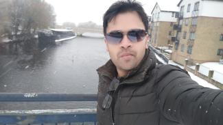 A man wearing sunglasses, standing on a bridge over a canal and looking at the camera