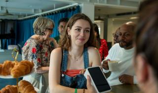 A young woman at a counter in a shop and smiling while using her phone to pay via a card reader held by somebody off screen.