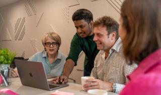 A man standing and demonstrating something on a laptop, watched by a man and a woman sitting either side.