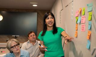 A woman standing beside a wall presenting to two people sitting beside her