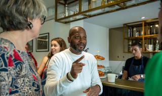 A man sitting in a coffee shop chatting to people standing around him and gesticulating with his hands
