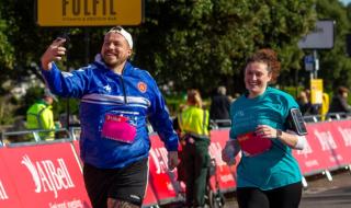 A man and woman running in a sponsored run. The man is holding a phone and taking a selfie