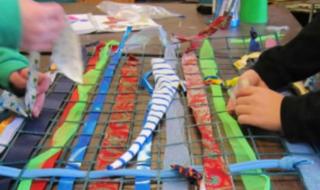 Two pairs of hands creating a mesh-style piece of art, involving strips of fabric and thread