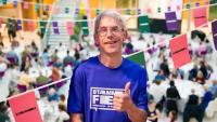 A man smiling and raising his thumb, aganist a backdrop of bunting and a crowded room