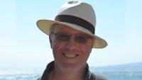 A man in a hat standing with the sea in the background, smiling for the camera