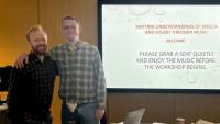 Two men standing beside a projection screen, with their arms around each other