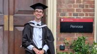 A young man in graduation robes, standing in front of a university door