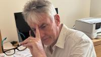 A man looking defeated, with a phone to his ear and his other hand holding a pair of glasses and touching his forehead