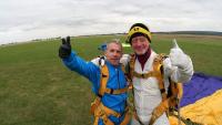 Two men in parachuting gear looking at the camera and smiling with arms around each other and their thumbs up