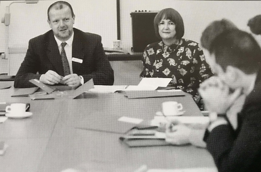A black and white image of a man and a woman at a desk speaking to people around it