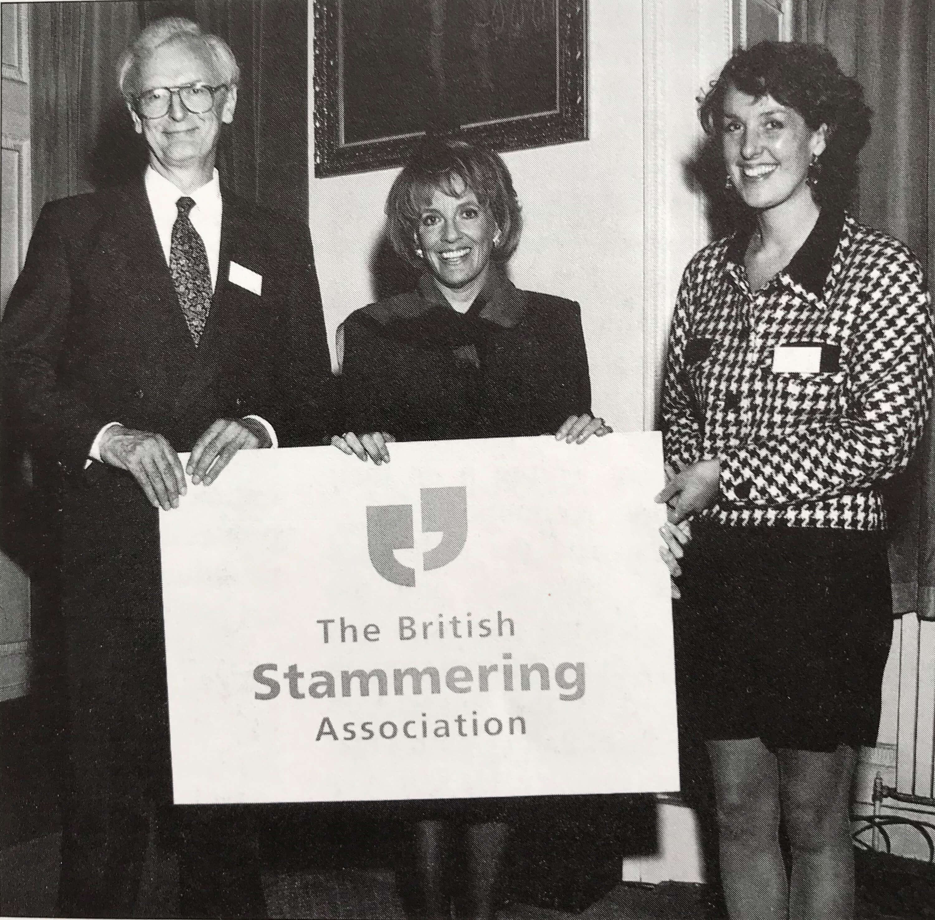 A black and white image of a man and two women holding a sign with the British Stammering Association logo on it