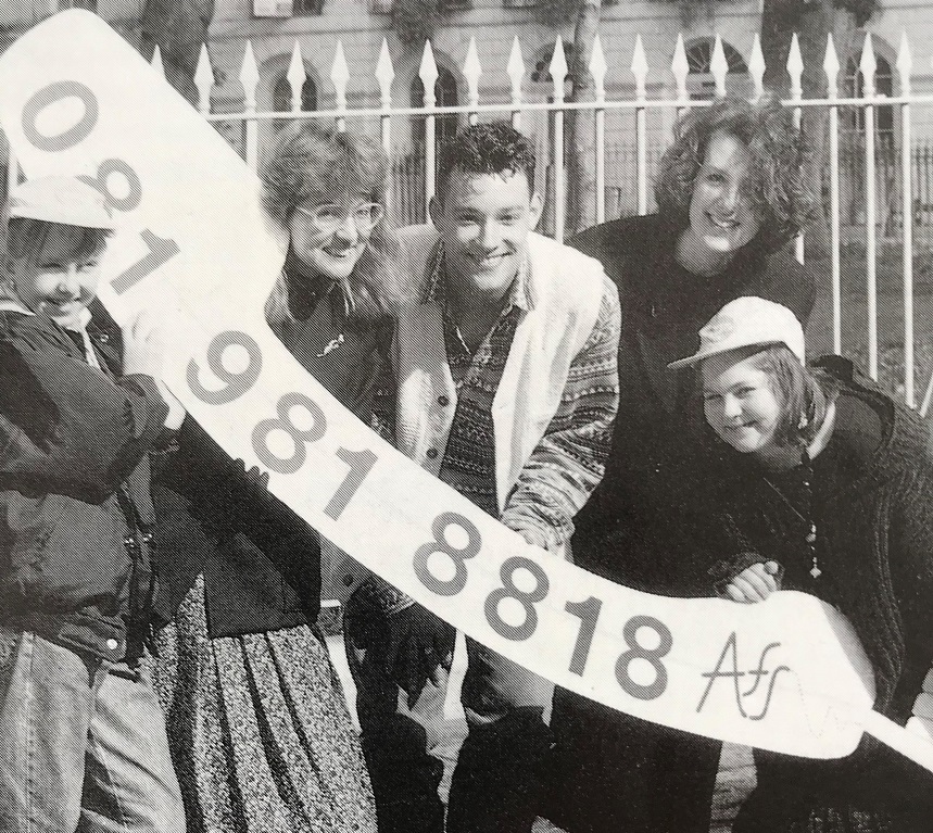 A black and white image of a group of people posing with a sash displaying a telephone number