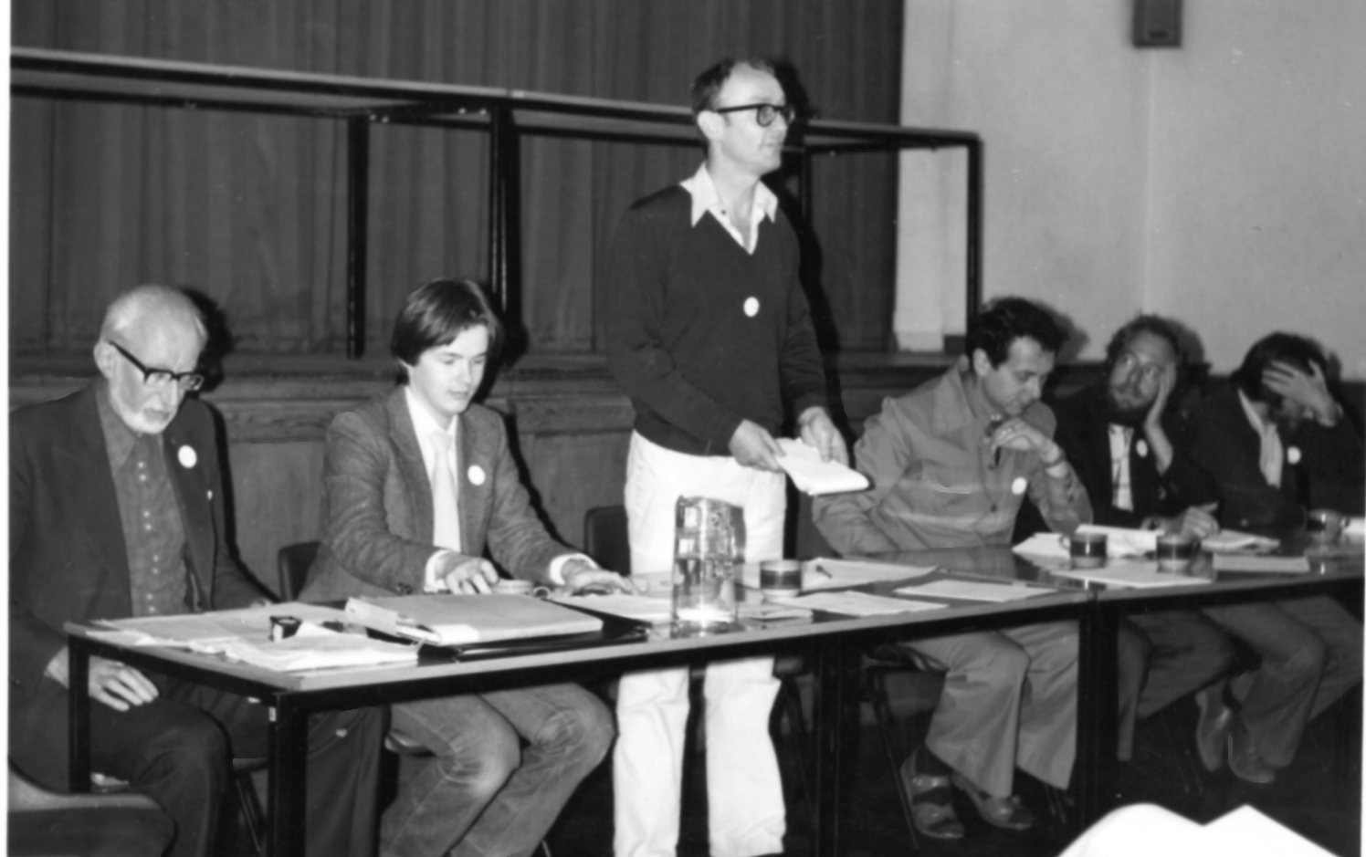 A black and white image of a board of trustees at a meeting, with one man standing and speaking