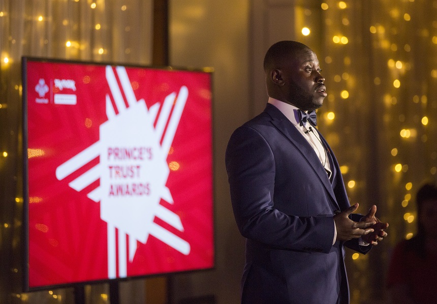 A man in a suit speaking to an audience in front of a screen with the text 'Prince's Trust Awards'